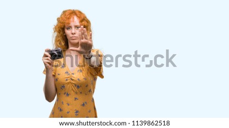 Young redhead woman taking pictures holding vintage camera with open hand doing stop sign with serious and confident expression, defense gesture