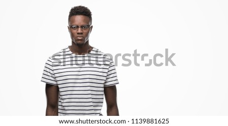 Young african american man wearing glasses and navy t-shirt with serious expression on face. Simple and natural looking at the camera.