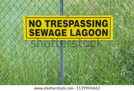lush wild green grasses behind a chain link fence with a yellow and black warning sign saying 'No Trespassing Sewage Lagoon'