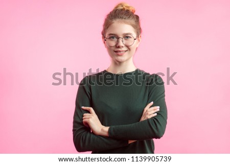 Attractive young woman in casual clothes wearing her hair in bun looking at camera with friendly charming smile, standing with crossed arms isolated over pink backgroind with copyspace.