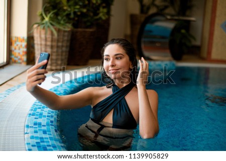 Young happy caucasian woman in fashionable swimmimg suite enjoying warm water and taking selfie in swimming pool at wellness center.