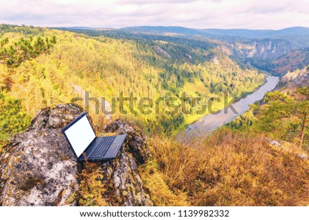 An outdoor laptop stands on a high-bank rock in a forest above the river.