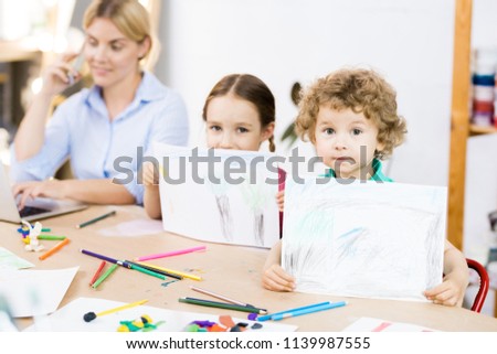 Portrait of two cute kids sitting at the table and showing their own pictures to the camera with teacher talking on phone in the background
