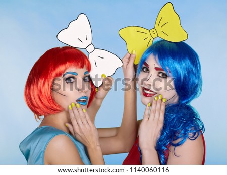 Portrait of young women in comic pop art make-up style. Females in red and blue wigs on blue background. Girls with yellow bow-tie in hands
