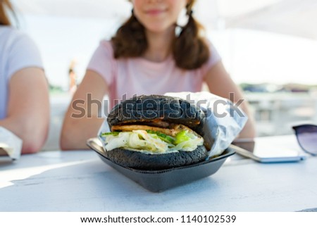 Close-up of black fast food burger. Background girls teenagers, outdoor summer cafe