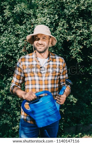 handsome male farmer in checkered shirt holding watering can and smiling at camera outdoors