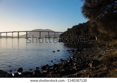 A beautiful sunset illuminates the silhouette of a tall bridge stretching over a calm river. The shoreline is protected by trees, and carpeted by rocks and pebbles.