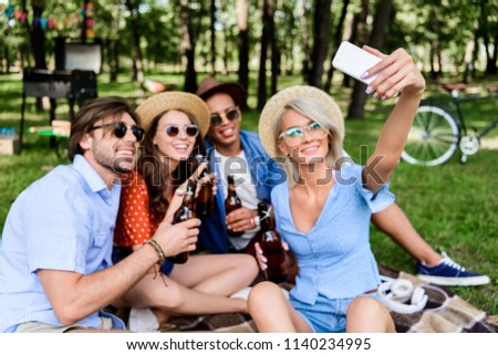 smiling multicultural friends with beer taking selfie on smartphone while resting in summer park