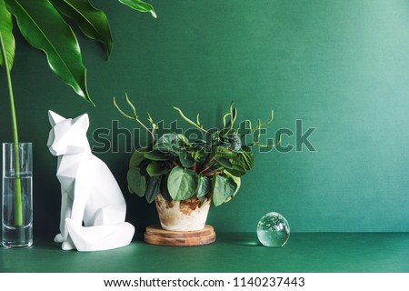 Green minimalistic room with cat figures, tropical leaf in glass vase and stylish plant. Design composition of desk in green interior.