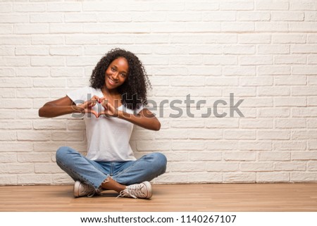 Young black woman sitting on a wooden floor making a heart with hands, expressing the concept of love and friendship, happy and smiling