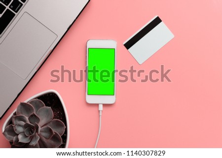 Top view of white smartphone with green chromakey on display. Flat lay with credit bank card, laptop, succulent plant on the light pink table as background.
