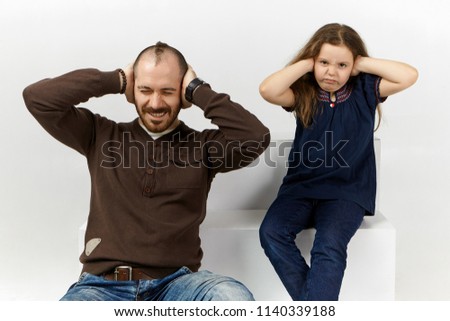 Picture of frustrated young unshaven male and annoyed ten year old female child posing in white studio covering ears with hands, irritated with annoying sounds, noise. Negative facial expressions