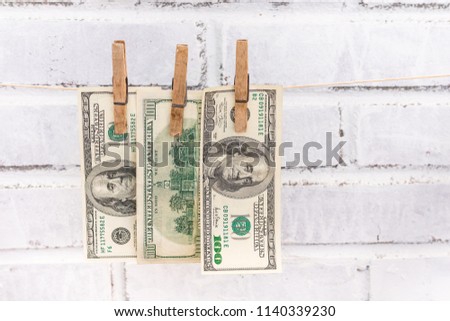 Three 100 USD bills hanging on the string attached with old wooden washing clips on the white shabby bricks background