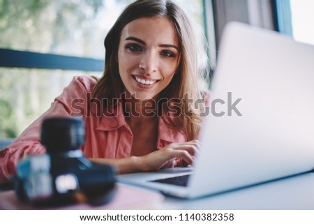 Portrait of cheerful attractive young woman working remotely at modern laptop computer using wireless high speed internet connection.Positive female photographer smiling at camera during distance job