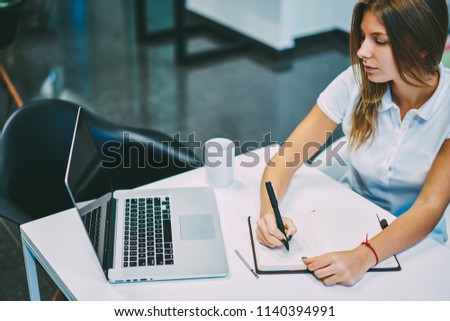 Pensive skilled student watching webinar online on laptop and making notes in notepad using wireless internet connection.Concentrated young woman making notes during reading information on web page