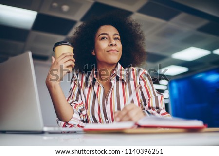 Pondering cute african american young woman writing down text information in notebook while thinking on creative ideas for publication.Contemplative dark skinned hipster student enjoying coffee