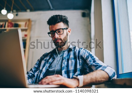 Concentrated male copywriter searching information for new article using modern technology and listening audio book via headphones, serious hipster guy in spectacles browsing internet on netbook