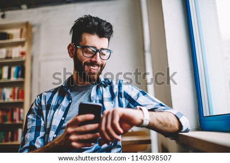Smiling successful hipster guy in eyewear checking time on wearable smartwatch while waiting for meeting at university campus, happy man holding mobile phone in hand and reading message on wristwatch Royalty-Free Stock Photo #1140398507