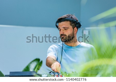 Photo of adult dj working with his equipment surrounded by palm leaves