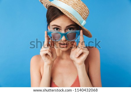 Portrait of a surprised young girl in swimsuit wearing sunglasses and summer hat posing isolated over blue background