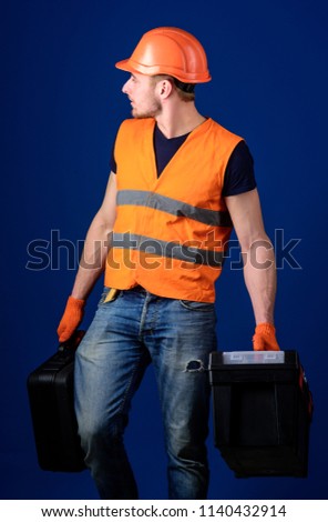 Man in helmet, hard hat holds toolbox and suitcase with tools, blue background. Equipped repairman concept. Worker, handyman, repairman, builder on calm face carries bags with professional tools.