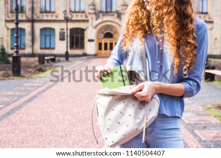 A female student with long curly hair takes a notebook from her backpack with a lecture summary in the courtyard of the university. Higher education concept