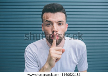Bearded man is looking on camera and showing the hush symbol. He is keeping finger on mouth. Isolated on striped and blue background.