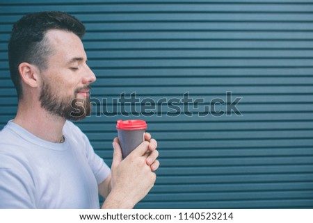 Nice and happy person is holding cup of coffee witht both hands. He is smiling. Guy is keeping eyes closed. Isolated on striped and blue background.