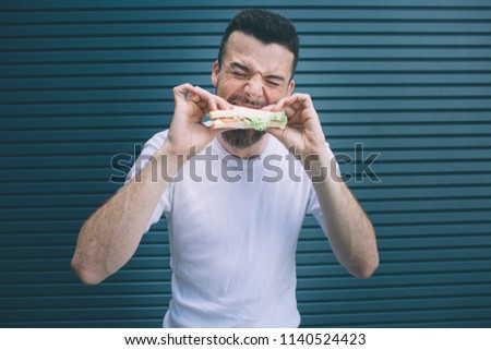 A picture of bearded man biting a piece of sandwich. He is holdng it with both hands. Guy is keeping eyes closed. Isolated on striped and blue background.