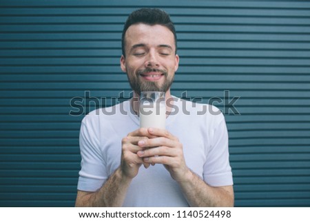 Portrait of person holding glass of milk close to face and enjoying it. He is keeping eues closed. Isolated on striped and blue background.
