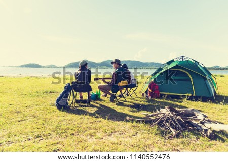 Group of travelers camping and doing picnic and playing music together. Mountain and lake background. People and lifestyle. Outdoors activity and leisure theme. Backpacker and Hiker. Back view angle