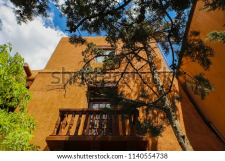 Perspective looking up at tall soutwestern stucco adobe building with balcony with corbels on a roof and trees framing the image under a blue sky with clouds