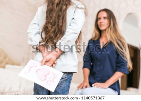 Charming little girl is giving her beautiful young mom a present, woman is smiling while sitting on couch at home