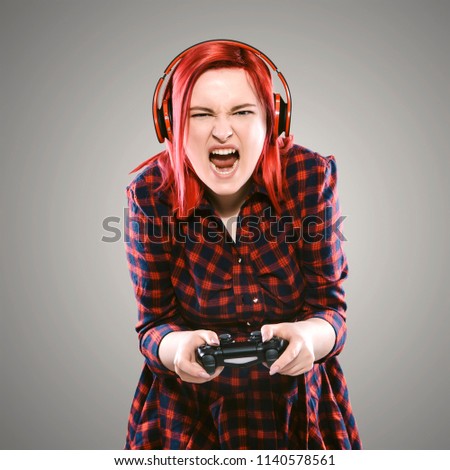 Red haired plus size ordinary woman standing on a neutral grey background. Emotional portrait. She playing in a game using joystick, wearing headphones