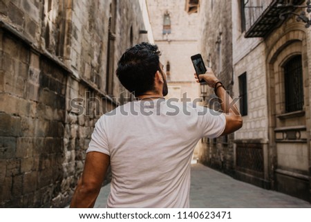 Content attractive hispanic man in white tshirt photographing himself on mobile phone while enjoying city travel.Bearded hipster male making selfie via smartphone on gotic streets