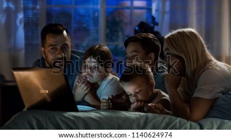 Excited parents with children chilling on bed at home in evening time and watching comic show on laptop all together