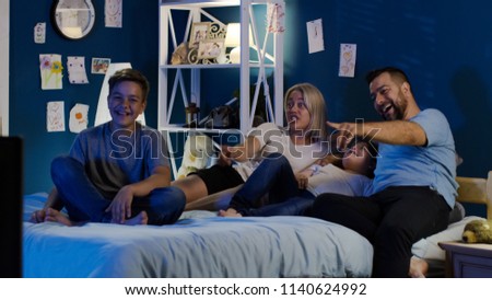 Adult parents with teen kids chilling on bed at night and watching comic show on TV laughing happily