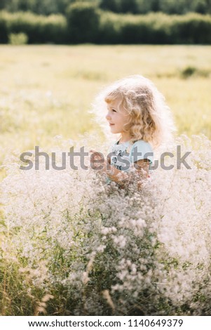 Adorable happy little girl with a curly blonde hair, wearing a light blue dress that says "flowers and hearts", standing in the sunny sunset field among wild grass and flowers, smiling