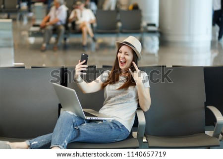 Young traveler tourist woman working on laptop doing selfie on mobile phone, show rock-n-roll sign waiting in lobby hall at airport. Passenger traveling abroad on weekends getaway. Air flight concept
