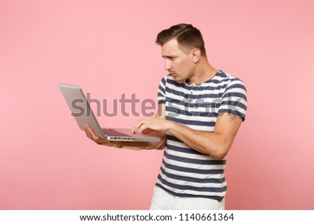 Portrait of concentrated young man in striped t-shirt working on laptop computer, copy space isolated on trending pastel pink background. People sincere emotions lifestyle concept. Advertising area
