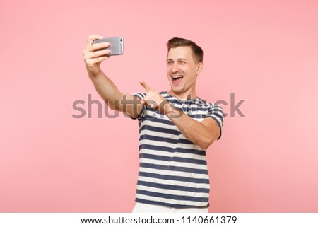 Portrait of smiling young man wearing striped t-shirt doing selfie shot on modern mobile phone isolated on trending pastel pink background. People sincere emotions lifestyle concept. Advertising area