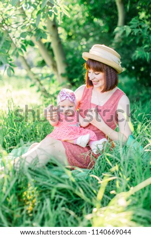 A happy family having a picnic in the green garden in a sunny spring day: a beautiful smiling mother sitting on green grass and her little laughing daughter on her legs