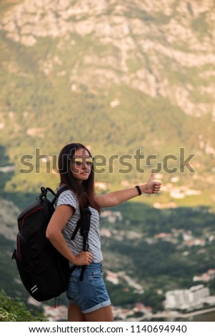 Young beautiful woman with black backpack hitchhiking standing on road. Beautiful young female hitchhiker by the road during vacation trip in mountains.