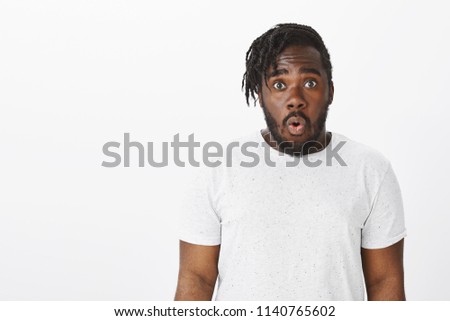Studio shot of shocked attractive plump dark-skinned guy in white t-shirt, saying wow with folded lips and opened mouth, staring with popped eyes at something surprising and amazing over grey wall