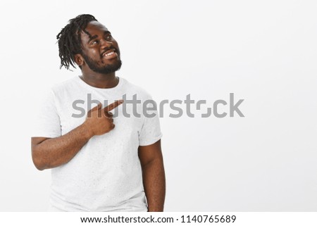 Studio shot of happy pleased attractive dark-skinned guy in white t-shirt, looking and pointing at upper right corner while smiling from satisfaction, seeing awesome copy space over background