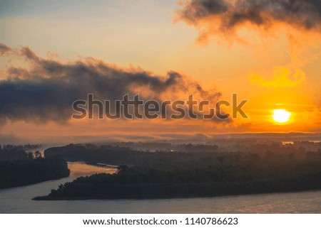 River flows along shore with forest. Channel flows around island in fog. Orange glow around sun at dawn in cloudy sky reflected on water. Morning atmospheric landscape of majestic nature in backlight.