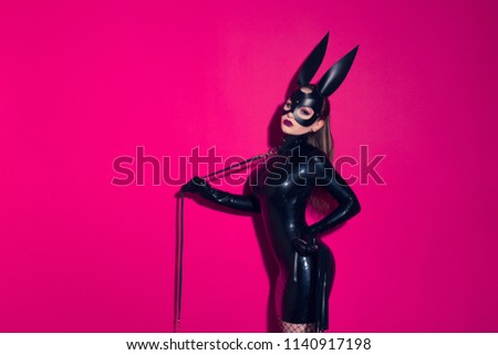 Beautiful dominant brunette vamp mistress bdsm girl with fashion makeup in glamour latex dress, collar and bdsm black leather fetish rabbit mask posing on hot pink background
