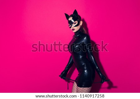 Beautiful dominant brunette vamp mistress bdsm girl with fashion makeup in glamour latex dress, collar and black leather fetish cat mask posing on hot pink background