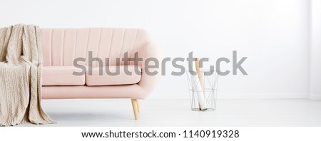 Real photo of white living room interior with metal basket with paper rolls and pastel pink couch with coverlet. Place for your armchair
