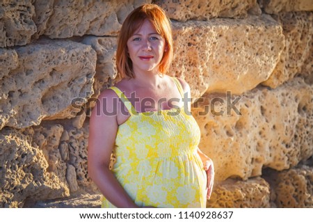 A portrait of a beautiful Caucasian pregnant woman in her early 30s in a yellow dress with an ancient limestone wall on her background.
